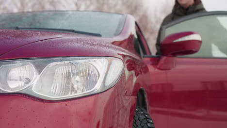 front angle view of red car with water droplets on headlight and hood during winter as young man in winter jacket enters vehicle, snowy background with bare trees enhances cold season atmosphere