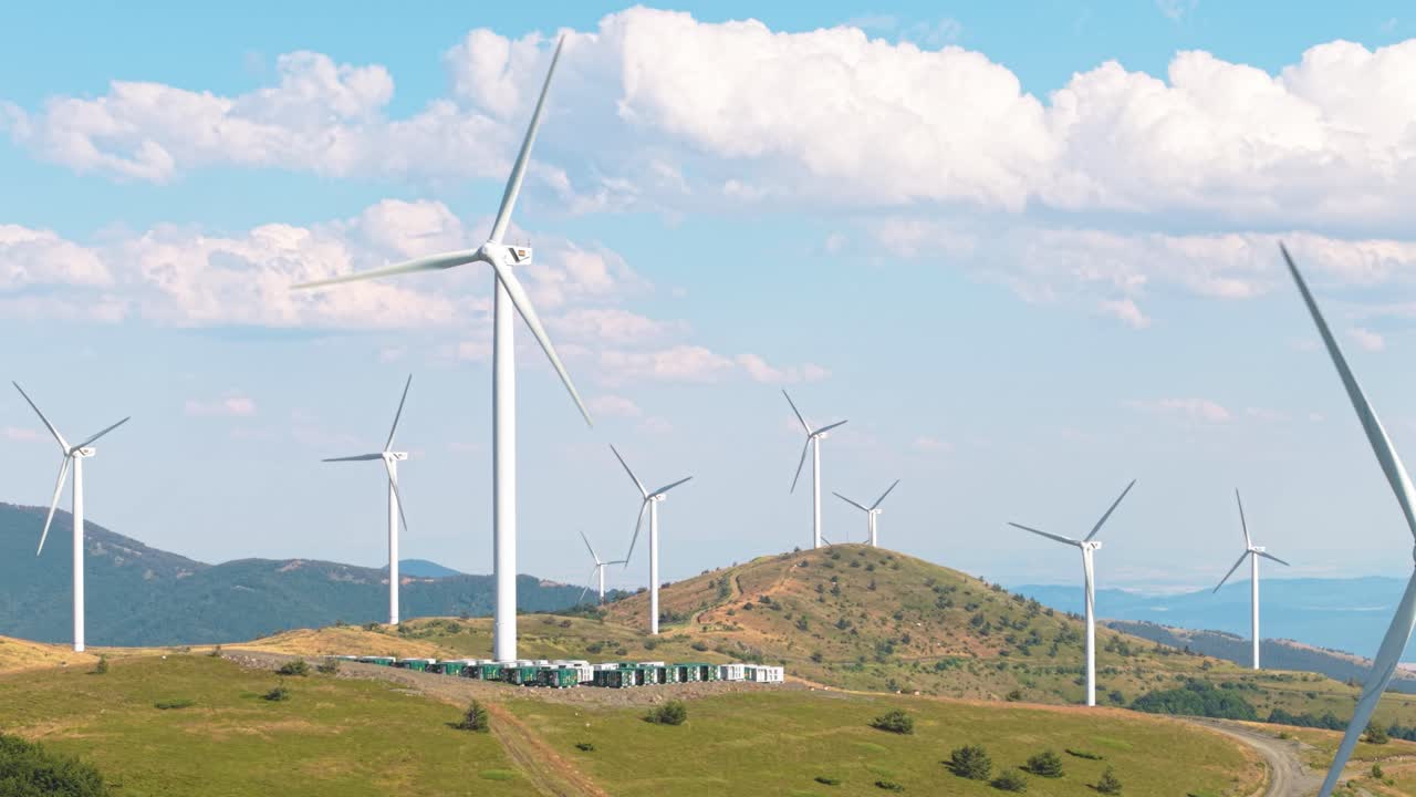 Drone footage of large wind turbines operating on elevated green terrain near Buzludzha. Scenic clouds and sky enhance clean power theme