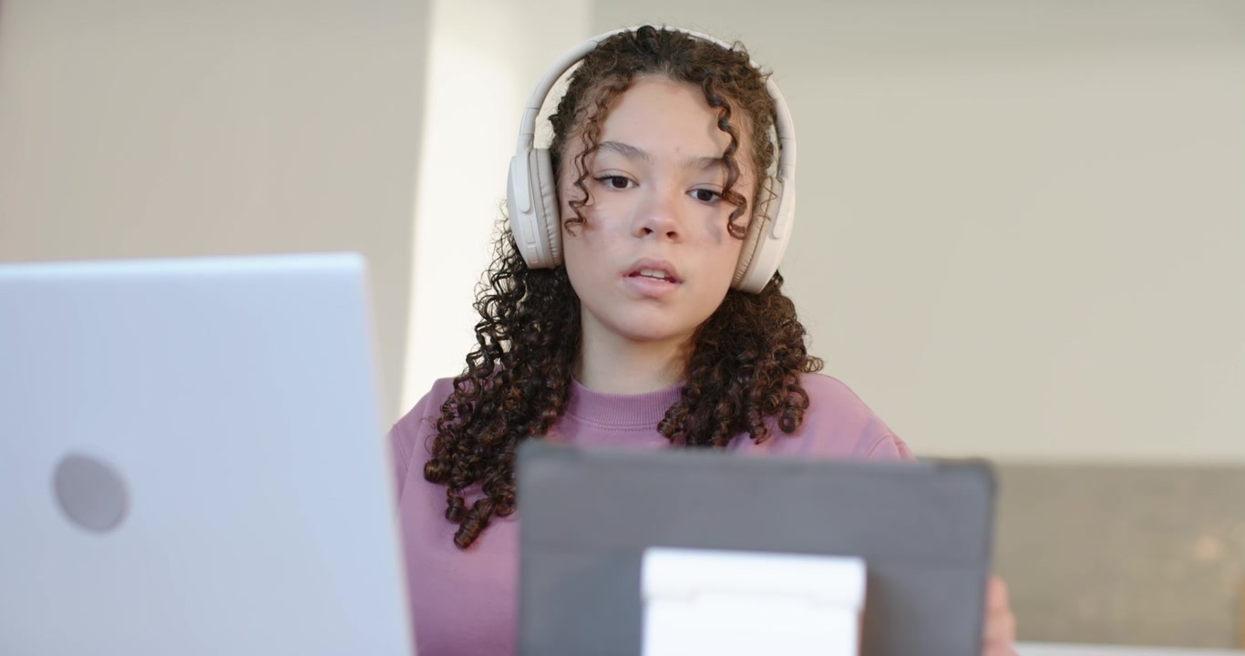 Afroamerikansk teenager i undervisning ved skrivebord, iført lilla top og headset, vekslende mellem bærbar computer, tablet og noter