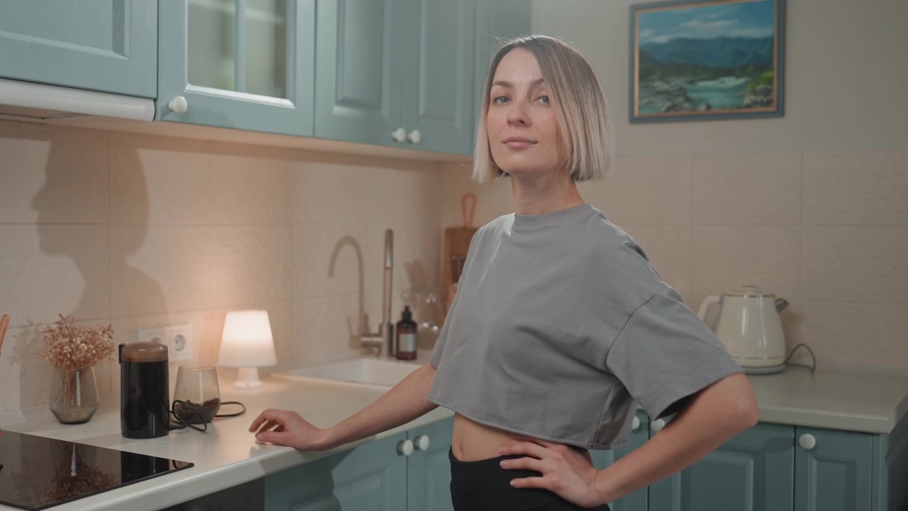 Cucina accogliente, giovane donna barista casalinga che prepara il caffè ascoltando musica, bancone illuminato da lampada, mobili color pastello, bollitore e macchina da caffè espresso sul bancone, mano sul fianco, caschetto biondo corto, sorriso rilassato, ambiente caldo