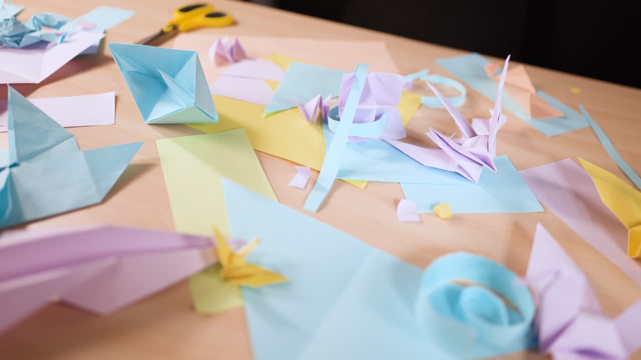 Una toma en ángulo alto muestra varias formas de origami en tonos pastel y restos de papel sobre un escritorio