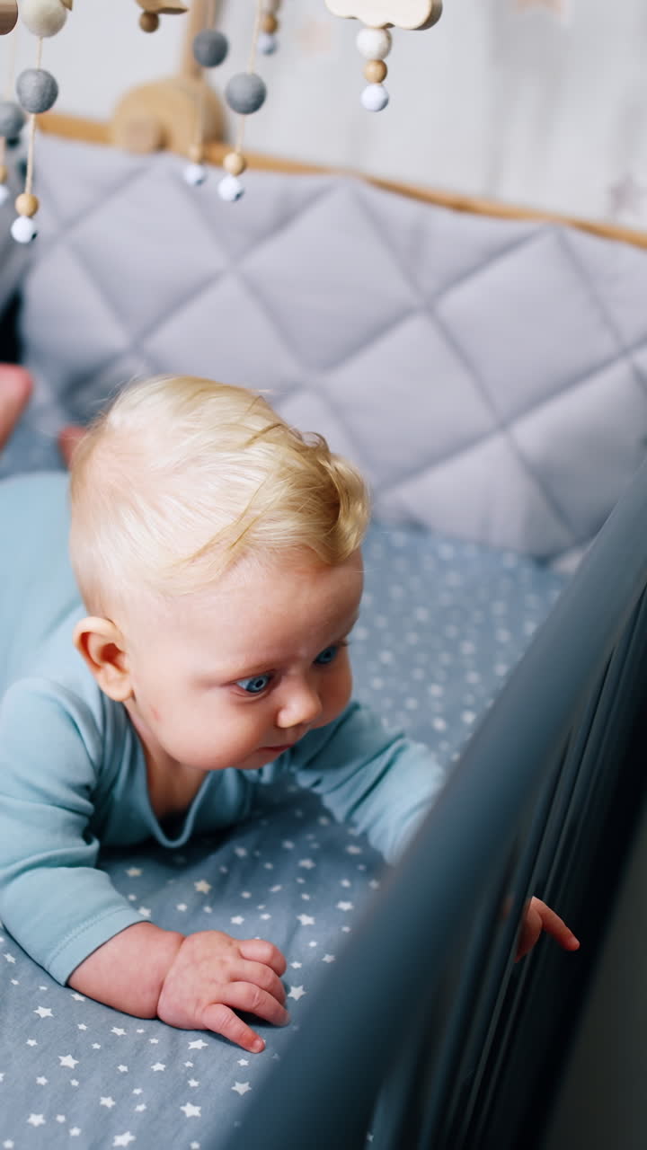Little cute child lying on the belly. Adorable infant baby in bodysuit lies in cot under the crib mobile. Vertical video