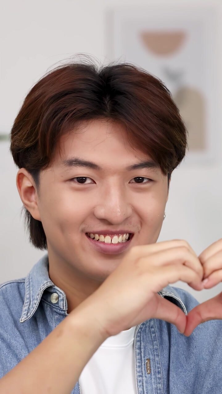 Un giovane uomo asiatico allegro con una camicia di jeans fa gesti a forma di cuore mentre sorride raggiante.