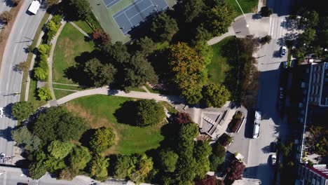 Höhenflug-über-Einem-Modernen,-Runden,-üppigen-Stadtpark-In-Einer-Luxuriösen-Wohngegend-In-Der-Innenstadt-Neben-Einem-Umweltfreundlichen-öffentlichen-Busservice,-Der-über-Die-Stadtstraßen-3-3-Mit-Elektrischen-Oberleitungslinien-Verbunden-Ist