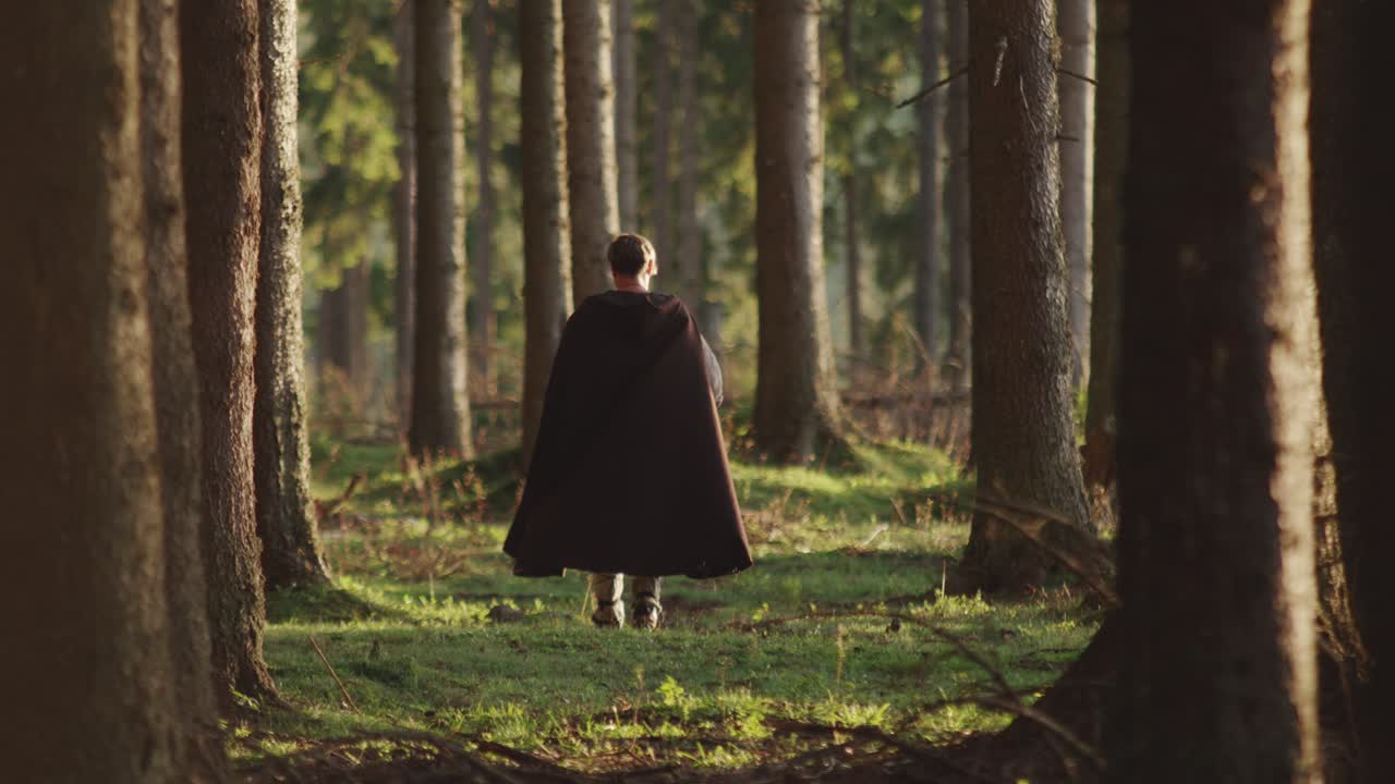 Cinematic shot of a historical or fantasy character in a dark cloak walking into a magical forest. Epic adventure, journey, or mystery concept for film and storytelling