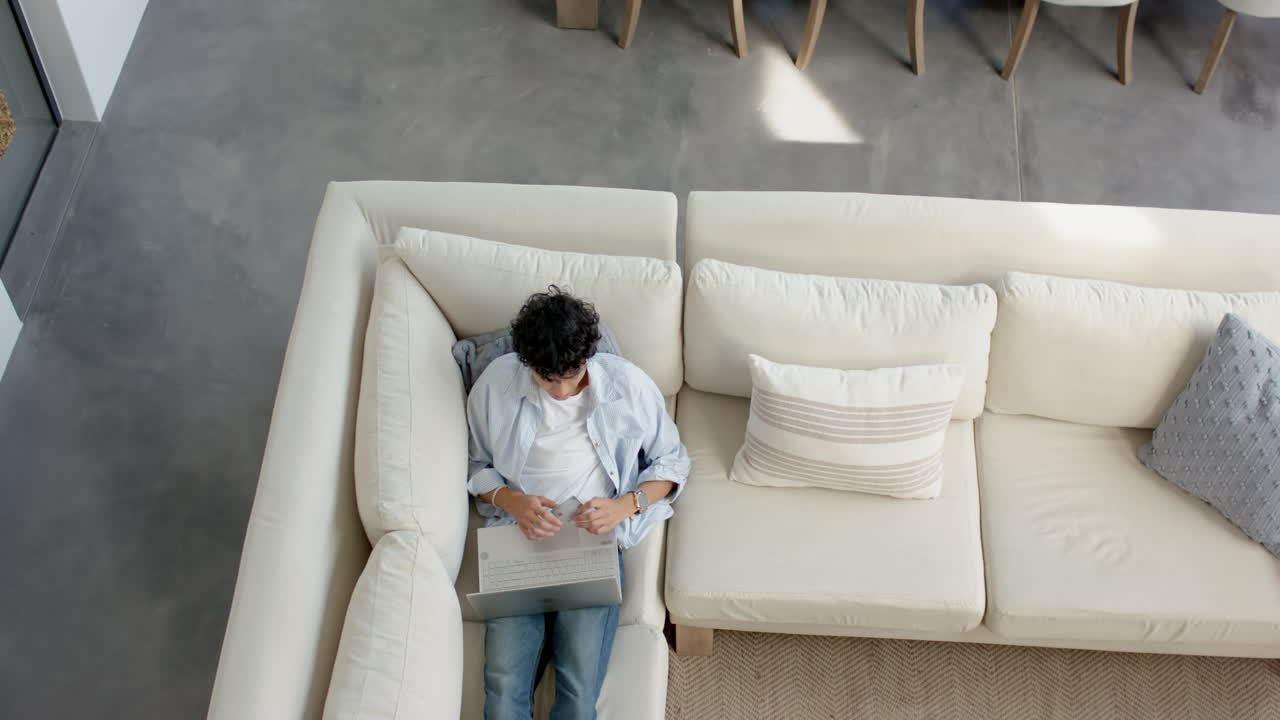 Using laptop, teenage boy relaxing on couch in modern living room, copy space