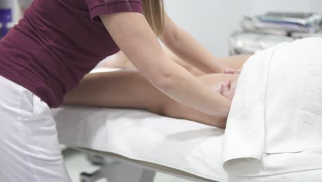 a professional physical therapy masseuse is massaging the posterior side of the leg of a young adult female caucasian client lying face down on a massage table, no face closeup with slow motion