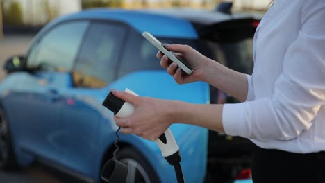 a woman is standing next to her electric car, charging it while using her cell phone. the vehicles hood is open, showing automotive parts like the tire and wheel