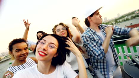 point of view shot of pretty girl holding camera and recording video of her dancing friends enjoying rooftop party. professional dj, mixing console and drinks are visible.