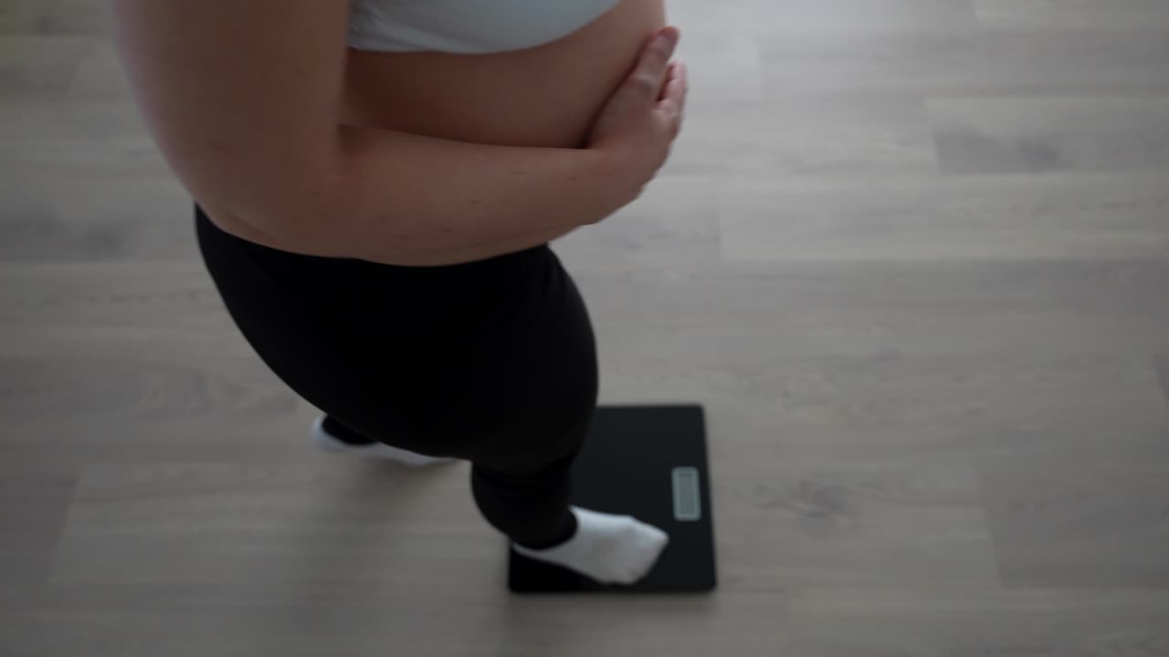 Pregnant woman checking weight while gently touching belly at home, third trimester moment