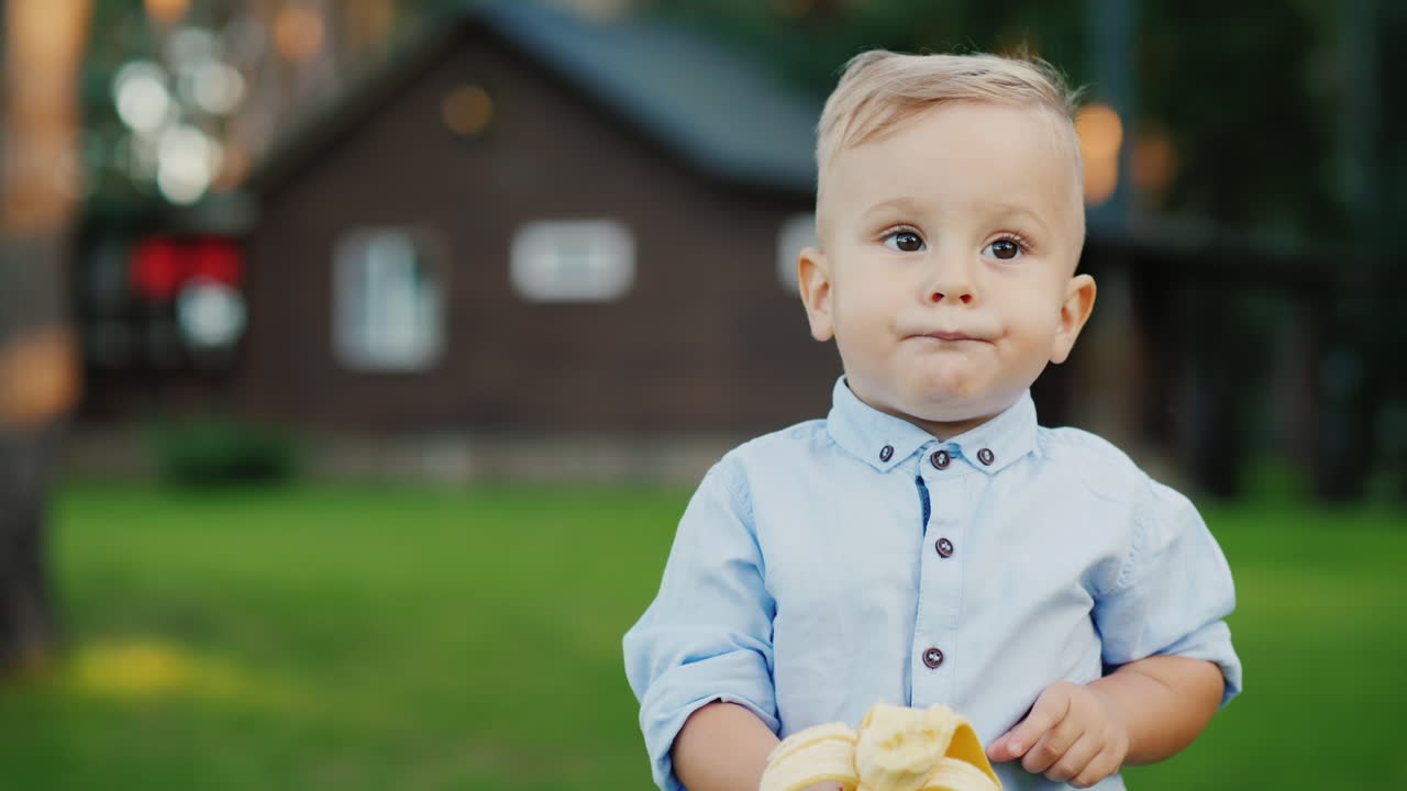 Cheerful 1 Year Old Baby Is Eating A Banana It's Standing On The Back Of The House 4k Video