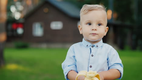 Fröhliches-1-jähriges-Baby-Isst-Eine-Banane,-Es-Steht-Auf-Der-Rückseite-Des-Hauses-4k-Video