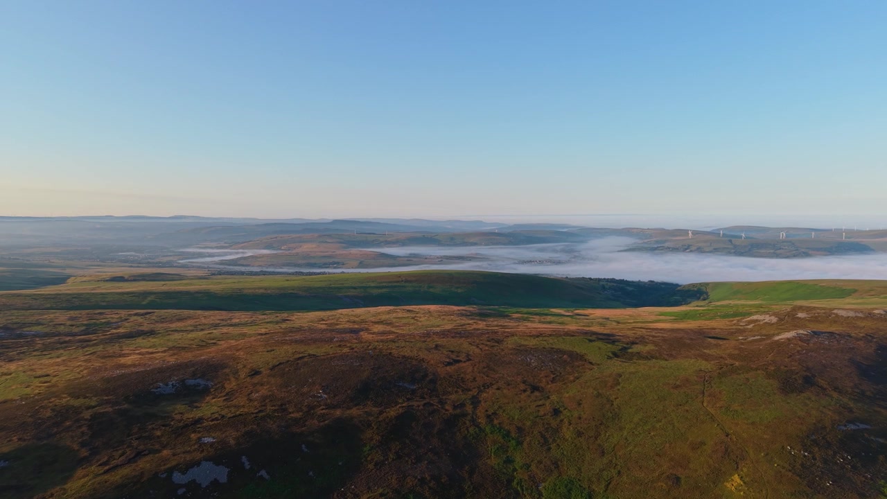 Vidstrakt naturlandskab i Sydwales med åbent hedelandskab og vindmøllepark i det fjerne. Tåge fylder dalene nedenfor