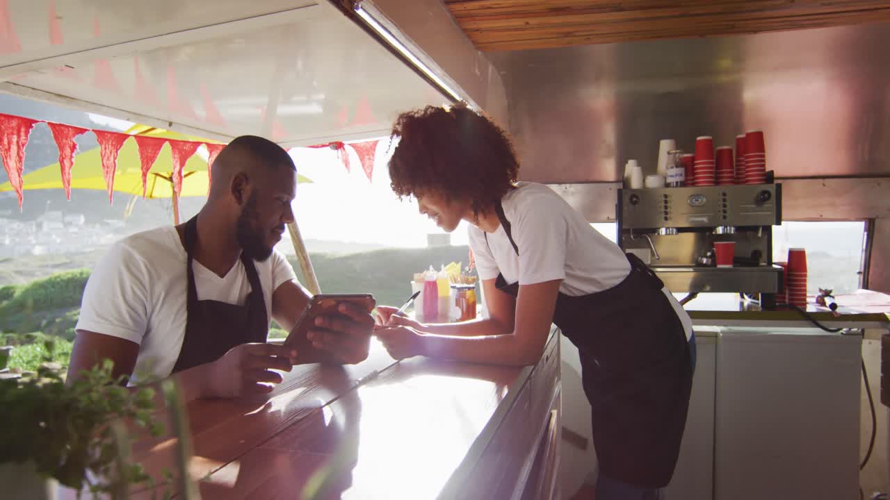 Afroamerikansk par iført forklæder bruger en digital tablet og tager noter i foodtrucken.