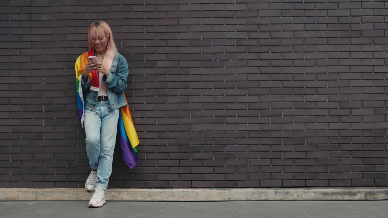 Woman using phone, leaning against brick wall, with rainbow flag