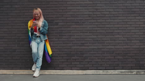 woman using phone, leaning against brick wall, with rainbow flag