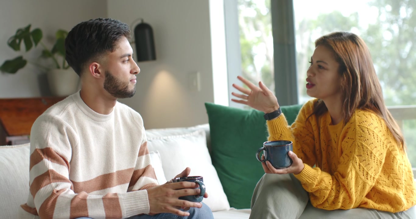 African American couple locking eyes, laughing, woman gesturing, man listening with mugs on couch