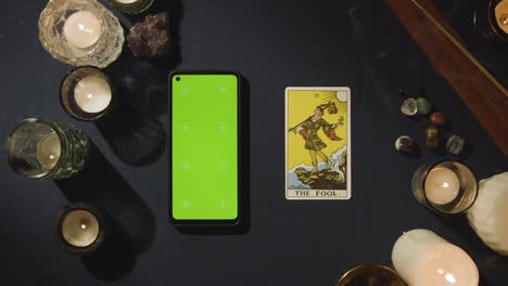 Fotografía-Cenital-De-Una-Persona-Que-Lee-Las-Cartas-Del-Tarot-Con-Un-Teléfono-Móvil-Con-Pantalla-Verde-Junto-A-La-Carta-Del-Tonto-En-La-Mesa.