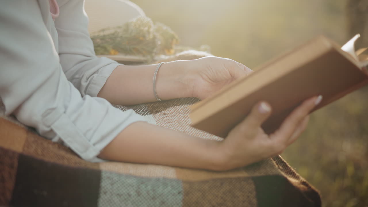 Primo piano di una mano con un braccialetto che sfoglia una pagina di un libro su una morbida coperta a quadri, la calda luce dorata del sole evidenzia i dettagli, creando un'atmosfera pacifica, accogliente e nostalgica.