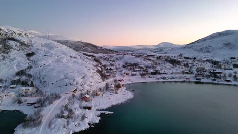 Flying-over-Ersfjordvegen-village-in-Norwegian-fjords-with-snowy-mountains,-houses-and-parked-boats