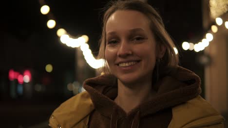 Una-Mujer-Joven-Y-Bonita-En-La-Calle-Con-Abrigo-Amarillo-Se-Vuelve-Hacia-La-Cámara-Sonriendo-Y-Brilla-Con-Destellos.-Luces-Detrás