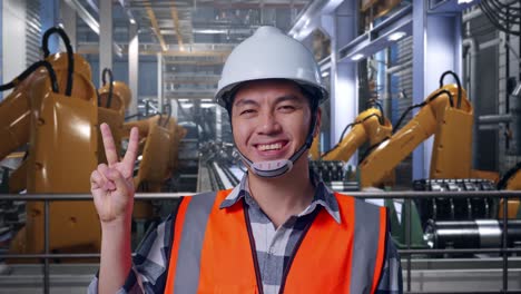 Close Up Of Asian Male Engineer With Safety Helmet Smiling And Showing Peace Gesture While Standing With Industrial Robotic Arms Working