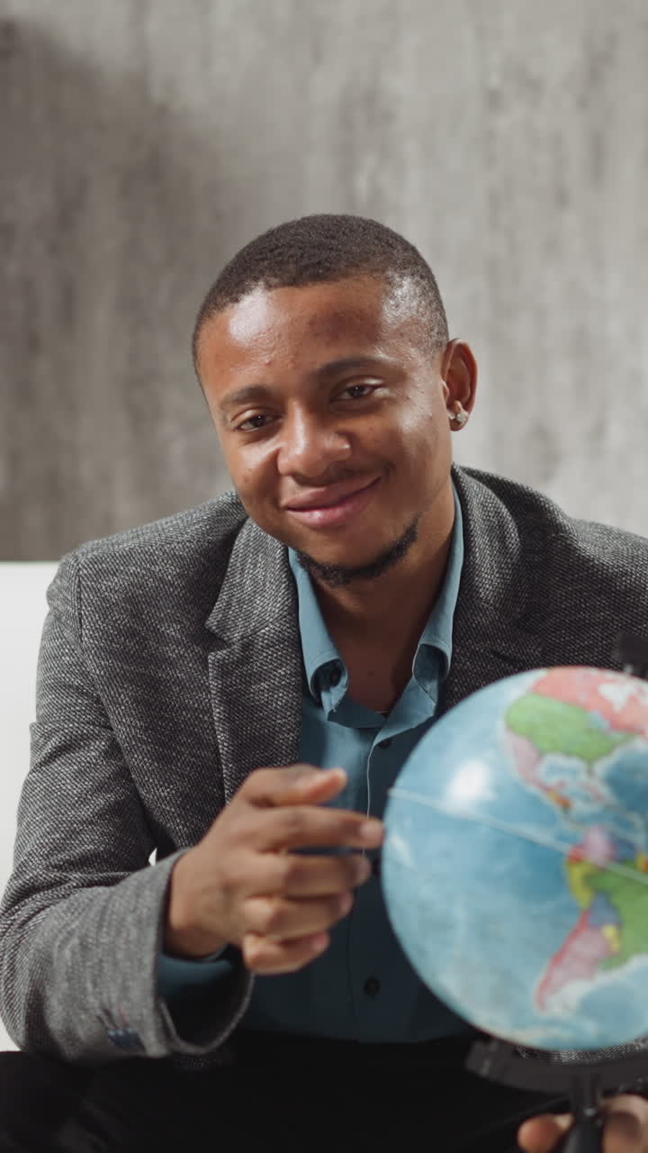 Smiling African-American geography teacher finger walks along globe inviting schoolchildren to travel at online lesson near laptop at home slow motion