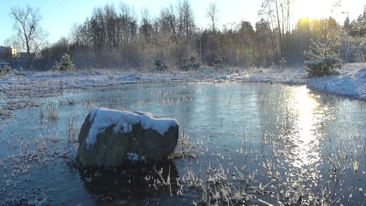 Langzaam bewegende beelden van een kleine bevroren, reflecterende vijver of een meer tijdens een zonnige winterdag, terwijl de zon ondergaat of opkomt boven het ijs. De natuurlijke grond en rotsen zijn bedekt met dikke, prachtige witte sneeuw.