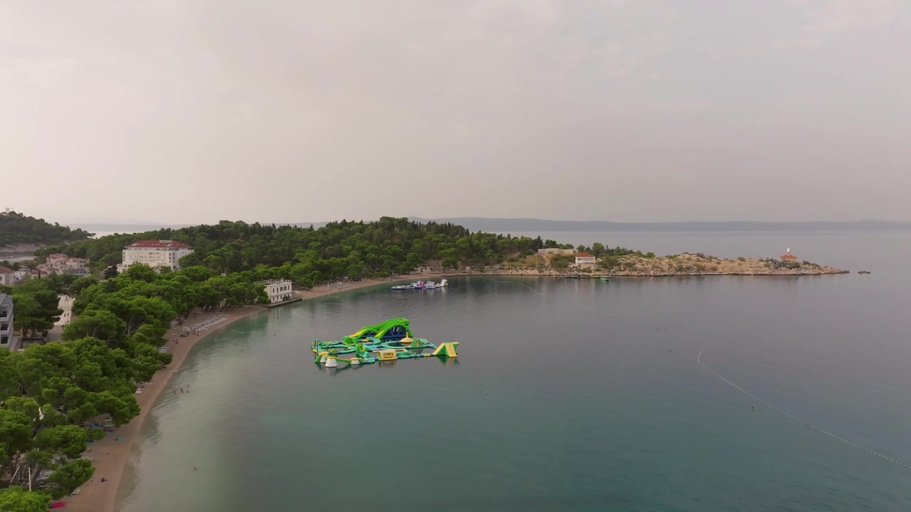 Parco acquatico gonfiabile che galleggia al largo di una tranquilla spiaggia alberata vicino a un piccolo promontorio a Makarska sotto un cielo coperto e soffuso
