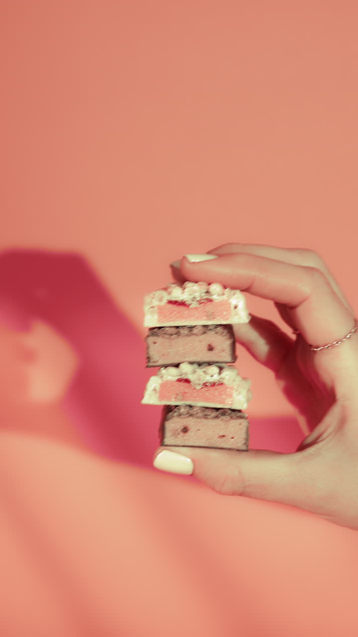 Close-up of a hand holding a neatly stacked four white strawberry and chocolate bars against a pink background. vibrant layers, crisp textures, soft lighting emphasize a clean, appetizing composition