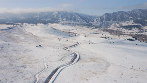 Aerial-moving-forward-to-reveal-hiking,-biking,-and-running-trail-covered-in-snow-that-leads-to-tall-Colorado-mountain-range-frozen-with-ice-and-covered-with-foggy-clouds-in-the-winter