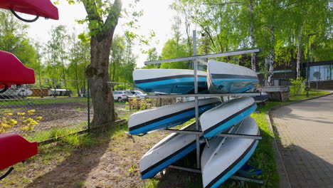 stack of blue and white kayaks stored on metal racks near a tree and a parking area, with red kayaks in the foreground