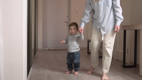 mother helps her baby to get up from the floor and take his first steps at home