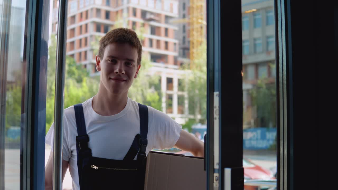 un mensajero con una camiseta blanca y un mono azul sostiene una caja en una mano, y con la otra abre la puerta de vidrio de un edificio moderno y entra. entrega de mercancías a la oficina