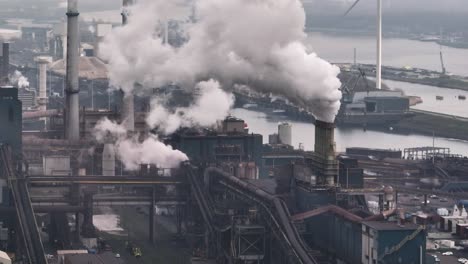 Aerial-panning-shot-of-Tata-steel-mill-in-the-port-of-Ijmuden-releasing-steam-and-pollution-with-the-blades-of-a-wind-turbine-cutting-through-the-frame