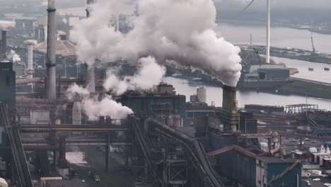 Aerial panning shot of Tata steel mill in the port of Ijmuden releasing steam and pollution with the blades of a wind turbine cutting through the frame