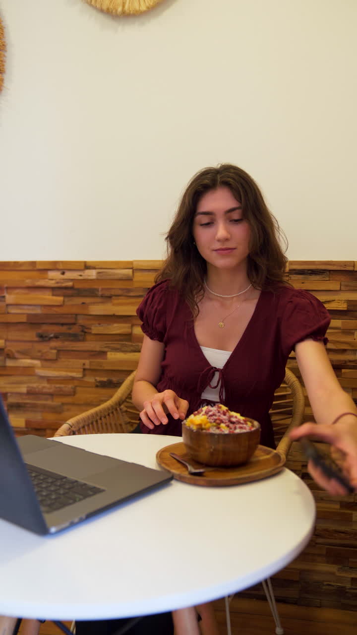 Young woman taking a selfie with her smartphone while holding an acai bowl and sitting at a table with a laptop in a cafe