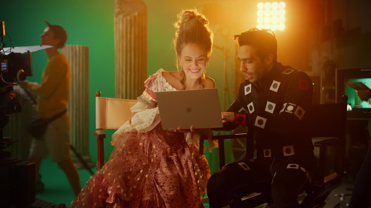 actriz sonriente con vestido renacentista y actor con traje de captura de movimiento sentado en sillas, usar computadora portátil. estudio de rodaje de película de alto presupuesto. en el estudio de cine período de traje de drama set de película