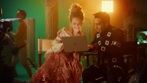 smiling actress wearing renaissance dress and actor wearing motion capture suit sitting on chairs, use laptop computer. studio shooting high budget movie. on film studio period costume drama film set