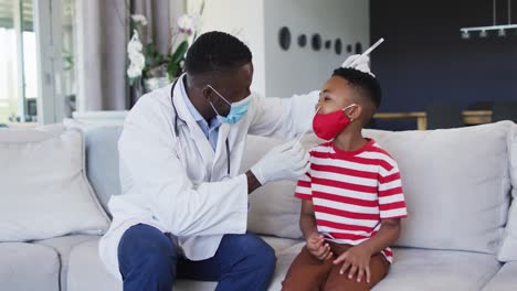 Afroamerikanischer-Arzt-Mit-Gesichtsmaske,-Der-Zu-Hause-Eine-Nasenabstrichprobe-Eines-Jungen-Entnimmt