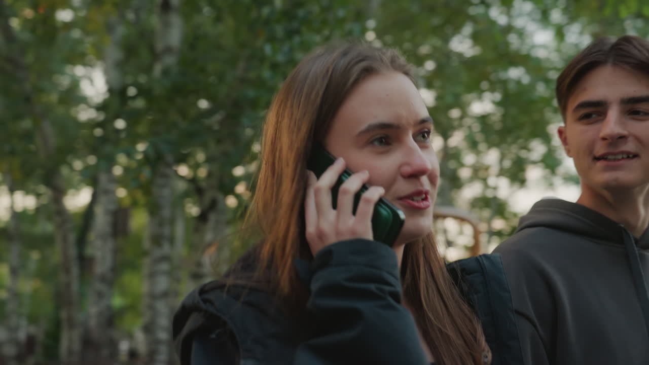 Mulher branca falando ao telefone ao ar livre enquanto um amigo branco se aproxima de moletom, reencontro espontâneo em caminho arborizado suburbano, sorrisos relaxados, luz do sol da tarde, interação jovem e casual durante uma caminhada