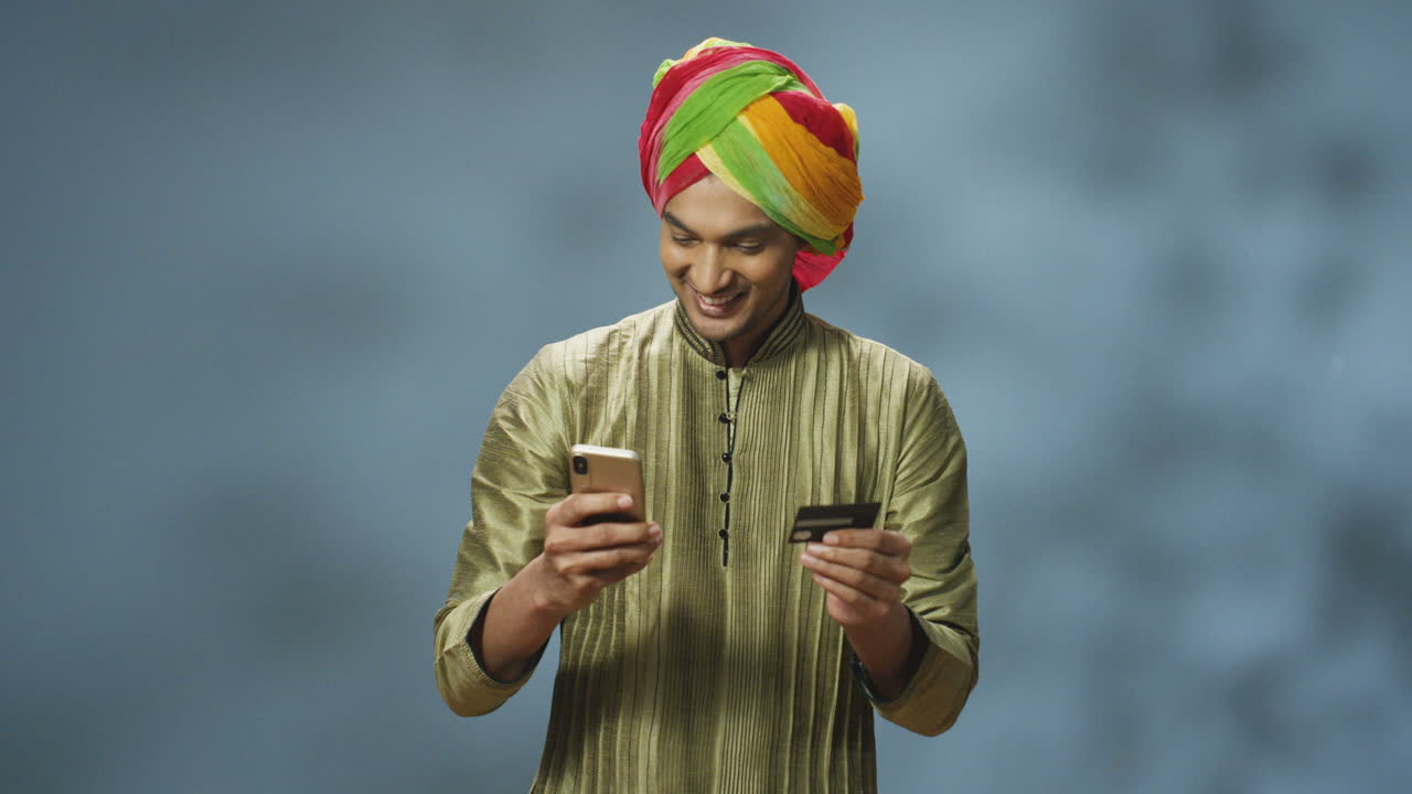 hombre indio guapo y alegre con turbante y ropa tradicional comprando en línea con un smartphone, luego se ríe y celebra