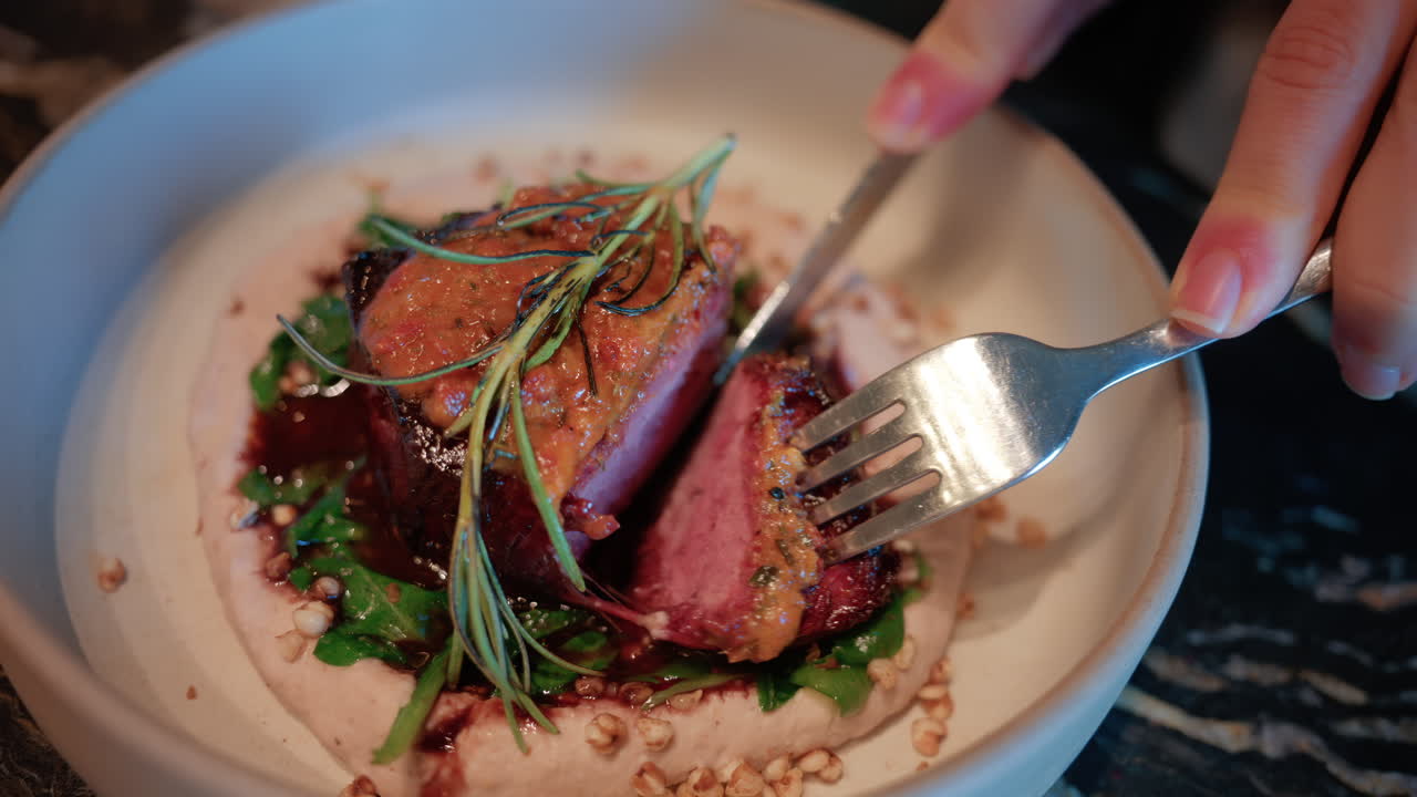Close up of a woman's hand cutting into a gourmet meat dish served with sauce, greens, and rosemary garnish on a white plate