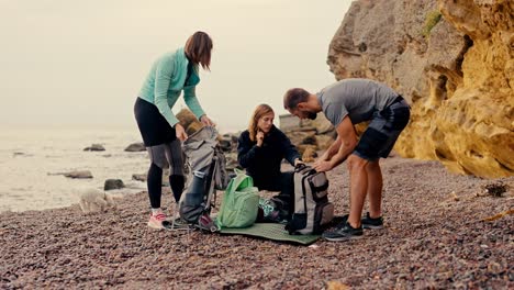 A-group-of-rock-climbers-unpack-their-backpacks-and-prepare-to-climb-the-yellow-rocks-near-the-sea-on-a-rocky-shore-in-summer.-A-blonde-girl-in-a-black-sports-uniform,-as-well-as-a-girl-with-a-bob-hairstyle-and-a-brunette-guy-are-taking-apart-their-backpacks-and-preparing-to-climb-the-rocks