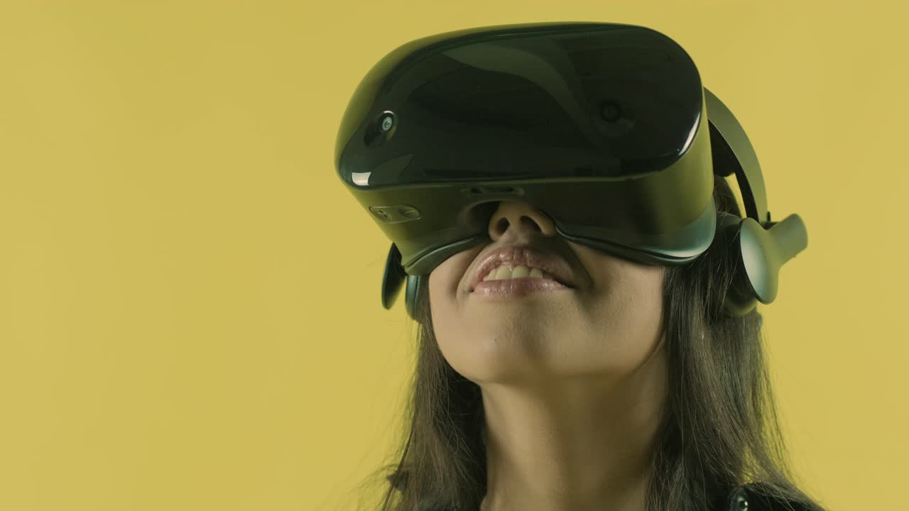 Young woman looks around and wonders how amazing  in the VR world. Virtual reality helmet on the yellow background. Portrait