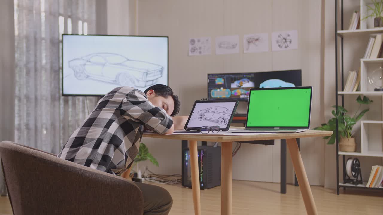 아시안 남성이 그림을 그리면서 잠을 자고 있는 모습: 테이블 에 녹색 스크린 노트북, tv와 컴퓨터를 탑재한 스튜디오에서 3d 전기 자동차 모델을 보여준다.
