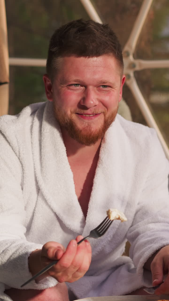 Man and wife enjoy breakfast together in cottage. Man tells story to woman eating pancakes at table with flowers. Romantic start of day at gampling resort