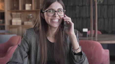 Charming-young-woman-in-a-glasses-is-sitting-in-a-cafe-and-talking-on-a-phone