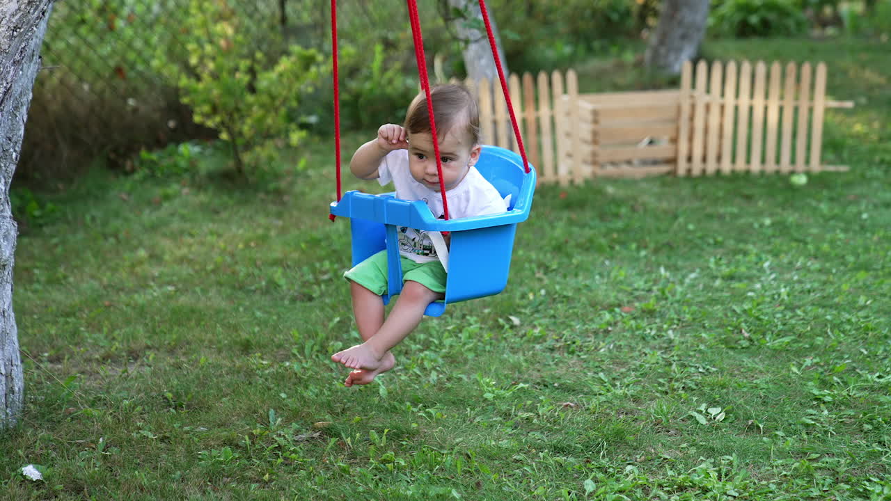 Little kid in t-shirt and shorts swaying in a swing. Calm barefoot baby boy in the garden in summer. Nature backdrop in blur.