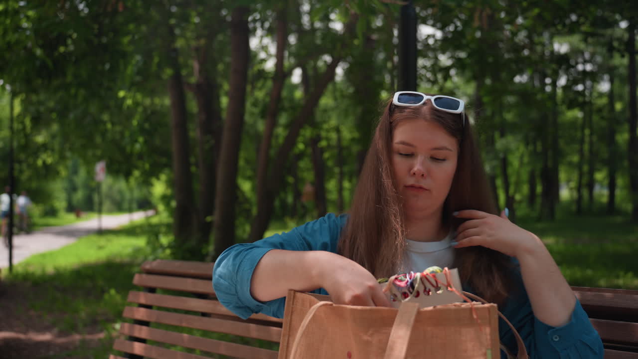 Young woman seated on wooden bench in green park carefully folding and placing checkered blanket back into brown tote bag, surrounded by trees under warm sunlight